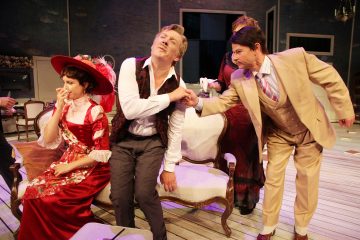 Lavalla esiintyy kolme näyttelijää värikkäissä aikakauden puvuissa. Punamekkoon ja hattuun pukeutunut nainen istuu sohvalla, liiviin pukeutunut mies nojaa dramaattisesti eteenpäin, ja toinen ruskehtavaan pukuun pukeutunut mies kumartaa ja suutelee hänen kättään.