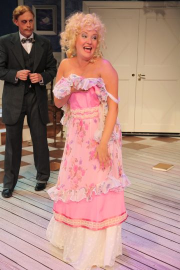 Nainen, jolla on vaalea kihara peruukki ja vaaleanpunainen kukkamekko, hymyilee kirkkaasti lavalla, kun taas pukuun ja rusettisolmioon pukeutunut mies seisoo hänen takkinsa takana ja säätää takkinsa. Kohtaus näyttää olevan osa teatteriesitystä.