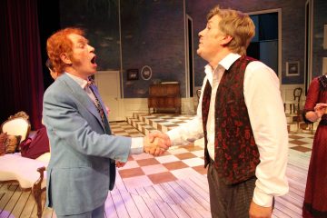 Kaksi pukumiestä kättelee lavalla, molemmat suu auki kuin puhuisivat tai laulaisivat. Lavasteet heidän takanaan muistuttavat vintage-olohuoneen kuviolattioita ja klassisia huonekaluja.