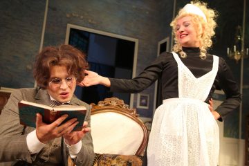 Silmälasipäinen henkilö lukee kirjaa keskittyneesti istuen tuolilla, kun hymyilevä, palvelijattareksi pukeutunut henkilö nykäisee leikkisästi hänen hiuksiaan teatteriympäristössä.