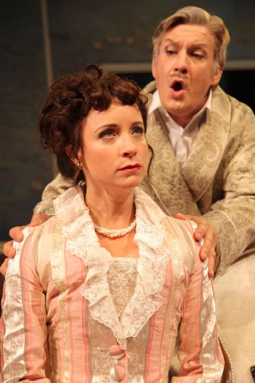 Pitsiseen, raidalliseen viktoriaaniseen mekkoon pukeutunut nainen istuu ilmeettömänä, kun hänen takanaan seisoo koristeelliseen kaapuun pukeutunut mies, joka puhuu painokkaasti kädet naisen olkapäillä.