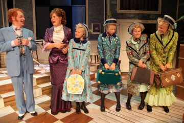 Lavalla seisoo joukko värikkäisiin, vanhanaikaisiin pukuihin pukeutuneita näyttelijöitä. Kaksi aikuista keskustelee vasemmalla, kun taas neljä naista, joilla on samanlaiset kukkamekot ja hatut, pitelee matkalaukkuja ja katselee toisiaan hymyillen teatteriympäristössä.