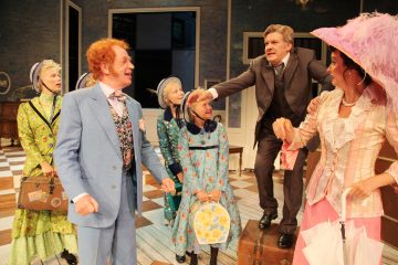 Ryhmä värikkäisiin aikakauden pukuihin pukeutuneita näyttelijöitä esiintyy lavalla. Yksi siniseen pukuun pukeutunut mies hymyilee, kun taas toinen ruskeaan pukuun pukeutunut mies seisoo rungon päällä ja osoittaa, kun kolme hattupäistä naista ja yksi vaaleanpunaiseen hattuun pukeutunut nainen katselevat.