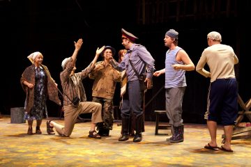 Ryhmä näyttelijöitä esiintyy lavalla puvuissa; yksi heistä polvistuu kädet kohotettuna, ja muut, muun muassa poliisin univormuun pukeutunut mies, reagoivat vilkkaasti hänen ympärillään kirkkaasti valaistussa lavastuksessa.