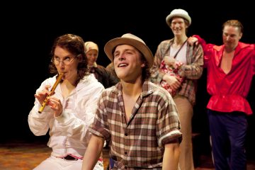 Lavalla viisi ihmistä, jotka ovat pukeutuneet värikkäisiin pukuihin. Edessä oleva henkilö soittaa nauhuria, kun taas toinen hattupäinen hymyilee vieressä. Kolme muuta seisoo taustalla nauraen ja vuorovaikutuksessa, mikä viittaa teatteriesitykseen.