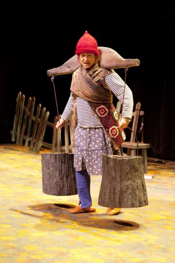 Värikkäisiin, epäsopiviin vaatteisiin ja punaiseen neulehattuun pukeutunut esiintyjä kantaa lavalla kaksi suurta puista ämpäriä ikeen varassa hartioidensa yli, ja taustalla on puisia rekvisiittaa ja aita.