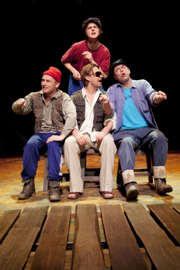 Neljä näyttelijää istuu ja seisoo näyttämöllä, pukeutuneina rentoihin ja karkeisiin vaatteisiin, ikään kuin soutaisivat venettä, ilmeikkäät kasvot, puiset lattialaudat edessä viittaavat laituriin. Tausta on tumma, mikä kiinnittää huomion esiintyjiin.