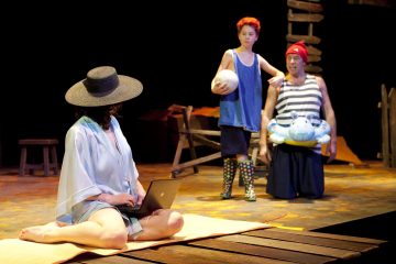 Aurinkohattuinen henkilö istuu pyyhkeellä kannettavan tietokoneen kanssa, kun taas lapsi pallon kanssa ja aikuinen, jolla on uimarengas ja punainen pipo, seisovat lähellä rantaa muistuttavalla lavasteella.