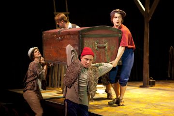 Neljä värikkäisiin pukuihin pukeutunutta näyttelijää nostaa ja kantaa suurta puista arkkua näyttämöllä teatteriesityksen aikana, ja taustalla näkyy rekvisiittaa ja tikkaat.