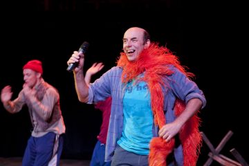 Lavalla esiintyy hymyilevä mies, jolla on kirkkaanoranssi höyhenboa ja mikrofoni kädessään, ja taustalla lavavalojen alla tanssii kaksi muuta ihmistä.