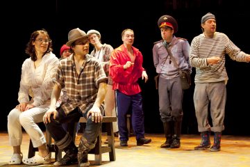 Ryhmä värikkäisiin pukuihin pukeutuneita näyttelijöitä esiintyy lavalla; jotkut istuvat ja toiset seisovat, mukaan lukien poliisin univormuun pukeutunut mies ja toinen raitapaitainen mies, mikä luo eloisan ja teatraalisen kohtauksen.