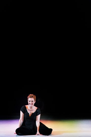 Lyhythiuksinen, mustaan mekkoon pukeutunut henkilö istuu hymyillen näyttämön lattialla valon valaisemana, ja hänen yläpuolellaan on suuri musta tyhjä tila.