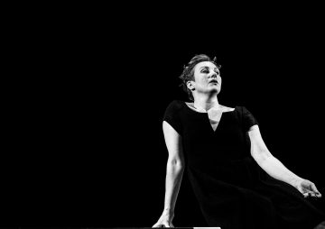 Tummapukuinen henkilö istuu mustaa taustaa vasten ja katsoo hieman ylöspäin mietteliäänä. Kuva on mustavalkoinen, mikä luo dramaattisen ja minimalistisen vaikutelman.