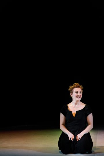 Mustapukuinen nainen istuu lattialla valonheittimen alla hymyillen ja hieman sivulle katsoen tummaa, tyhjää taustaa vasten.