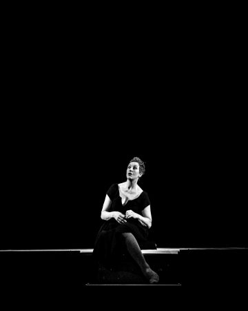Tummapukuinen nainen istuu yksin matalalla korokkeella mustaa taustaa vasten, jota valaisee yksi valo, mikä luo dramaattisen ja mietiskelevän tunnelman.