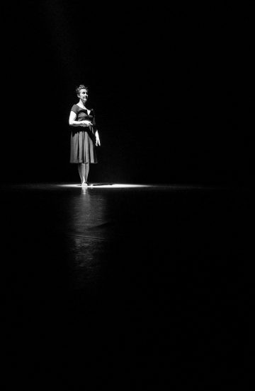 Tummapukuinen nainen seisoo yksin valokeilassa muuten pimeällä näyttämöllä, mikä luo dramaattisen ja teatraalisen tunnelman.