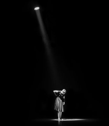 Tummapukuinen nainen seisoo yksin lavalla yhden valonheittimen alla, katsoo alaspäin toinen käsi lantiollaan, pimeyden ympäröimänä.