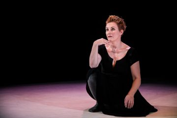 Lyhyet, punertavat hiukset omaava nainen, jolla on musta mekko, polvistuu hämärästi valaistulla näyttämöllä ja katsoo mietteliäästi nostamalla kätensä leukaansa. Tausta on tumma ja lavan lattia on pehmeästi valaistu.