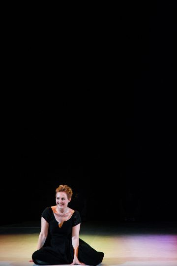 Mustaan mekkoon pukeutunut nainen istuu hymyillen lavan lattialla, tumma tausta ja lämpimät valonheittimet valaisevat häntä.