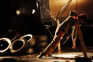 Henkilö nojaa sivuttain puulankulle lavalla, käsi roikkuu alaspäin, ja häntä ympäröivät pinotut autonrenkaat ja dramaattinen valaistus teatteriympäristössä.