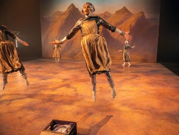 Metallipukuihin ja kuulokkeisiin pukeutuneet esiintyjät hyppivät ilmassa lavalla, jonka taustalla on vuoristo. Etualalla hiekanvärisellä lattialla on pieni avoin laatikko.