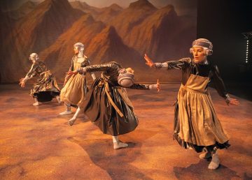 Neljä esiintyjää metallisissa puvuissa ja päähineissä tanssii lavalla maalatun vuoristotaustan edessä ja ottaa ilmeikkäitä asentoja kädet ojennettuina ja taivutettuina luoden liikkeen ja draaman tunteen.