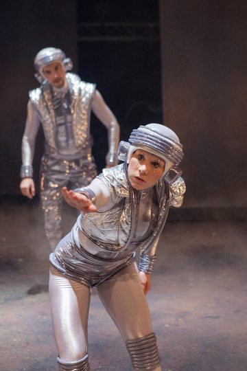 Lavalla on kaksi ihmistä, joilla on hopeanväriset futuristiset puvut ja kypärät. Edessä oleva henkilö kurottautuu eteenpäin ilmeikkäällä eleellä, kun taas takana oleva henkilö seisoo hieman epäselvästi. Tausta on tumma ja sumuinen.