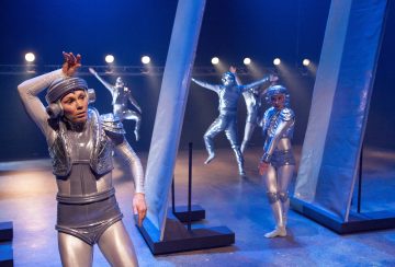 Neljä esiintyjää futuristisissa, hopeisissa puvuissa poseeraa lavalla, jossa on geometriset lavasteet ja dramaattinen valaistus. Yksi on lähellä kameraa käsi kohotettuna, kun taas muut poseeraavat tai liikkuvat taustalla.