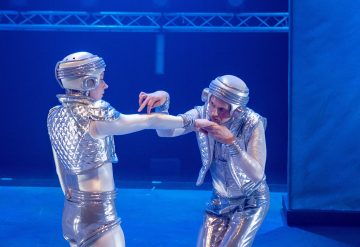 Kaksi hopeanhohtoisiin futuristisiin pukuihin ja kypärään pukeutunutta henkilöä esiintyy lavalla sinisessä valaistuksessa. Toinen ojentaa kätensä ja toinen kumartuu suutelemaan kättään, molemmat näyttelevät dramaattisesti.