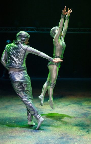 Lavalla on kaksi esiintyjää, joilla on hopeanväriset, futuristiset puvut ja kypärät. Toinen hyppii kädet kohotettuna, kun taas toinen seisoo takana ja ojentaa kätensä. Valaistus heittää näyttämölle vihertävää hehkua.