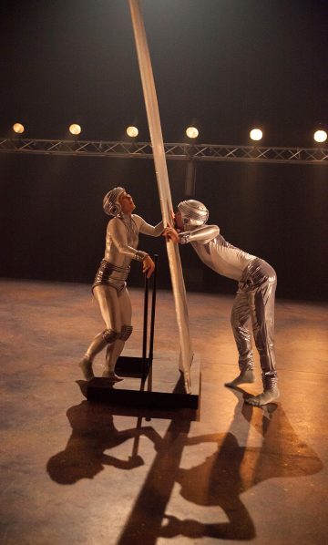 Kaksi esiintyjää kiiltävissä hopeisissa puvuissa ja kypärissä ovat vuorovaikutuksessa suuren pystysuoran paneelin kanssa lavalla lämpimissä näyttämövaloissa, toinen seisoo ja toinen nojaa eteenpäin paneelia vasten.