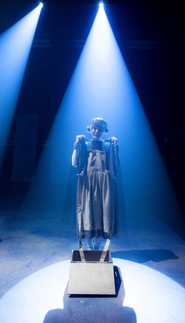 Henkilö seisoo laatikolla dramaattisen sinisen näyttämövalaistuksen alla ja pitää mekkoa edessään molemmin käsin, kun hänellä on kuulokkeet ja otsalamppu.