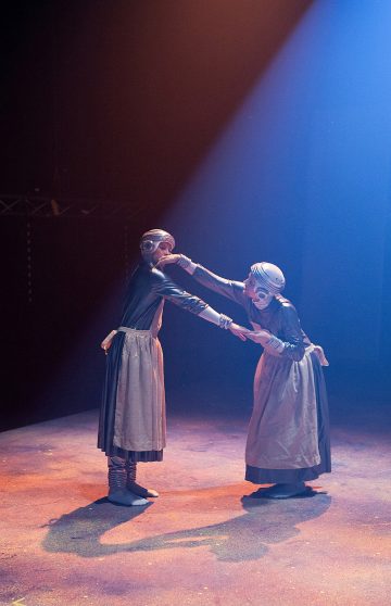 Kaksi pitkiin hameisiin, esiliinoihin ja metallikypäriin pukeutunutta esiintyjää kohtaa toisensa valonheittimen alla, kurottautuu ja koskettaa toistensa kasvoja dramaattisessa, hämärästi valaistussa näyttämökohtauksessa.