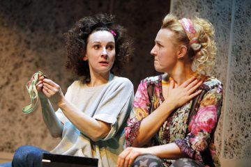 Kaksi naista istuu tiiviisti vierekkäin; toinen, jolla on tummat kiharat hiukset, pitää raidallista narua kädessään ja näyttää yllättyneeltä, kun taas toinen, jolla on vaaleat kiharat hiukset ja kukkavaatteet, pitää kättään rinnallaan ja vaikuttaa tunteelliselta.