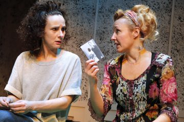 Kaksi naista istuu lähekkäin, toisella on harmaa paita ja huolestunut ilme, ja toinen, jolla on kukkapusero, näyttää hänelle vilkkaasti mustavalkoista valokuvaa. Kohtaus viittaa intensiiviseen keskusteluun.