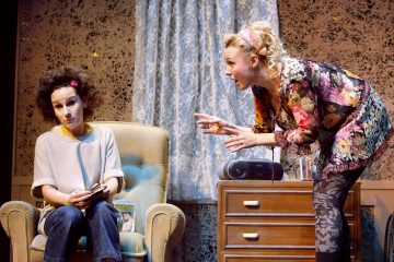 Kaksi naista huoneessa: toinen istuu nojatuolissa vaikuttuneen näköisenä puhelin kädessään, toinen seisoo vieressä ja puhuu vilkkaasti juoma kädessään. Taustalla on lipasto, pitsiverhot ja radio.