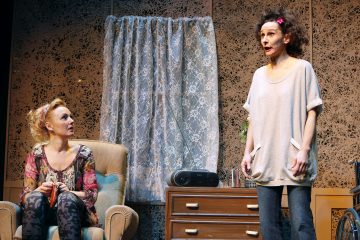 Kaksi naista on huoneessa, jossa on kuviolliset seinät ja pitsiverho. Toinen nainen istuu nojatuolissa ja katsoo ylöspäin, kun taas toinen nainen seisoo ja puhuu vilkkaasti. Taustalla näkyy radio ja lipasto.