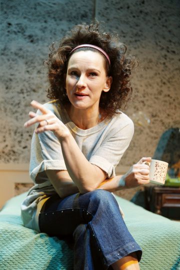 Nainen, jolla on kiharat hiukset ja vaaleanpunainen otsanauha, istuu sängyllä, pitää toisessa kädessään kahvikuppia ja elehtii toisella kädellään. Hänellä on harmaa paita ja siniset farkut. Taustalla on pehmeästi valaistu pilkullinen seinä.