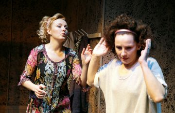 Lavalla on kaksi naista. Toinen vaaleatukkainen ja kukkamekkoinen nainen elehtii ilmeikkäästi, kun taas toinen kiharahiuksinen ja hiuspantainen nainen nostaa kätensä puolustautuvasti ja näyttää ahdistuneelta. Taustalla on vaateteline ja kuvioitu seinä.