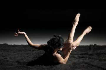 Mustaan asuun ja päähineeseen pukeutunut henkilö poseeraa tummalla, kuvioidulla pinnalla jalat koholla ja kädet ojennettuina sirosti dramaattisessa valaistuksessa mustaa taustaa vasten.