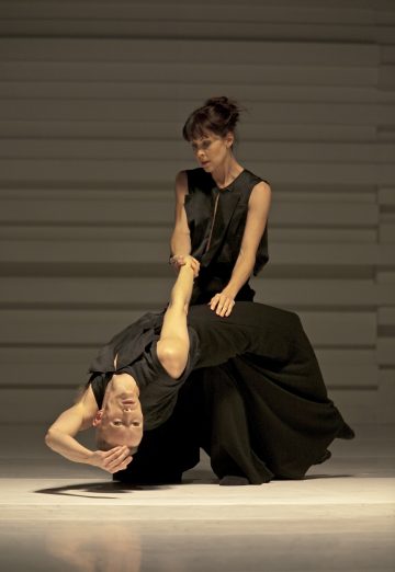 Kaksi mustiin asuihin pukeutunutta tanssijaa esiintyy minimalistisella lavalla. Toinen tanssija kumartuu taaksepäin toisen otteen tukemana, pää lähellä lattiaa ja käsi koholla, kun taas toinen tanssija seisoo pystyasennossa takanaan.