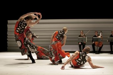 Neljä tanssijaa, jotka ovat pukeutuneet elinvoimaisiin oransseihin ja mustiin pukuihin, esittävät näyttämöllä ilmeikkäitä ja dynaamisia liikkeitä, kun taas kolme muusikkoa jousisoittimiensa kanssa istuu taustalla ja soittaa musiikkia. Tausta on minimaalinen ja vaalea.