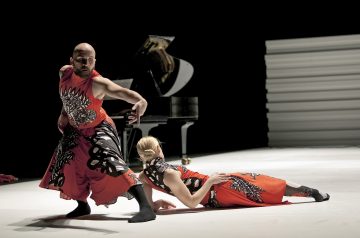 Kaksi nykytanssijaa punaisissa ja mustissa kuvioiduissa puvuissa esiintyy valkoisella näyttämöllä; toinen tanssija seisoo dramaattisessa asennossa ja toinen makaa lattialla. Taustalla näkyy flyygeli ja valkoinen tausta.