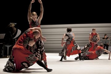 Lavalla esiintyy viisi tanssijaa, joilla on samanlaiset punaiset ja mustat puvut, ja kukin heistä ottaa ilmeikkäitä ja dynaamisia asentoja. Taustalla näkyy piano ja kaksi muusikkoa minimalistista lavastusta vasten.