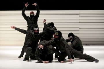 Kuusi mustiin asuihin ja rohkeasti kuvioituihin naamareihin pukeutunutta tanssijaa poseeraa dramaattisesti valkoisella näyttämöllä, jonka taustalla on valkoinen, harjakuvioinen tausta. Heidän liikkeensä ovat ilmeikkäitä, kädet ojennettuina ja vartalot vääntyneinä erilaisiin dynaamisiin asentoihin.