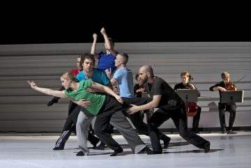 Ryhmä tanssijoita esittää lavalla nykytanssirutiinin dynaamisissa asennoissa ja ojennetuin käsin, kun muusikot taustalla soittavat jousisoittimia. Lavatausta on valkoinen ja minimalistinen.