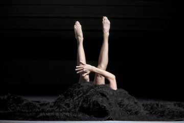 Kaksi paljasta jalkaa ja ristissä olevat käsivarret nousevat ylöspäin suuren, mustan, karvaisen peitteen alta tummalla näyttämöllä, ja muu vartalo jää kankaan peittoon.