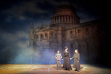 Lavalla seisoo kolme näyttelijää aikakauden puvuissa, kaksi lasta ja aikuinen nainen kädestä pitäen. Taustalla on kuvitettu kupolirakennus, joka tuo mieleen historiallisen tai teatterin ympäristön. Lämmin valo korostaa kolmikkoa.