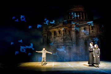 Lapsi seisoo kädet ojennettuina hämärästi valaistulla näyttämöllä, ja ympärille heijastetaan sinisten lintujen siluetteja. Kaksi tummiin vaatteisiin pukeutunutta aikuista katselee sivusta suuren, varjoisen kuvan edessä, joka esittää klassista kupolirakennusta.