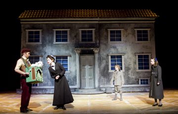 Neljä näyttelijää esiintyy näyttämöllä suuren, harmaan, kaksikerroksisen talon lavastuksen edessä; yksi näyttelijä pitää kädessään vihreää laatikkoa, jossa on valkoinen esine, ja muut katselevat 1900-luvun alun pukuihin pukeutuneina.